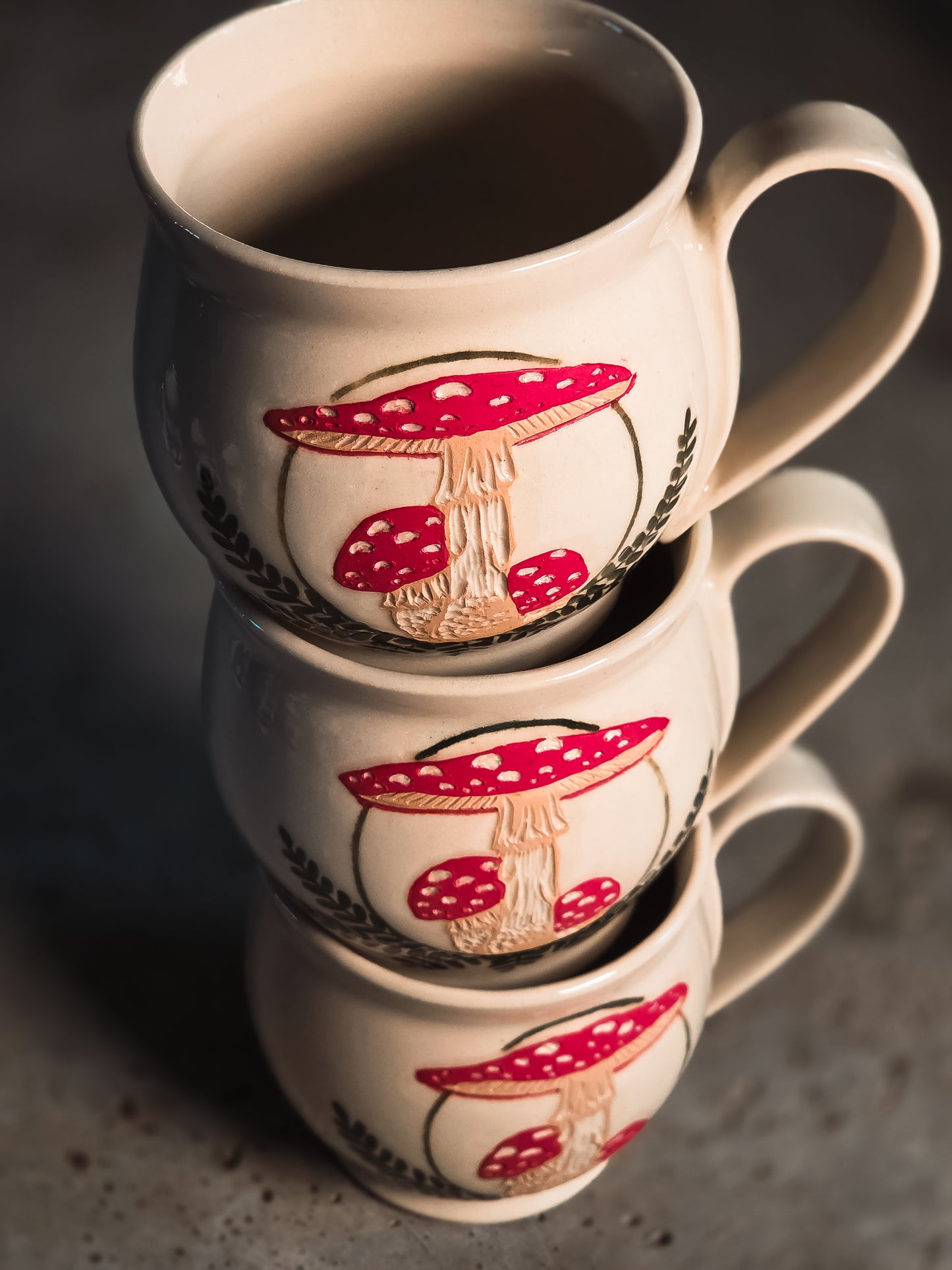 Cream ceramic mugs stacked, decorated with vibrant red and white Amanita mushroom designs.