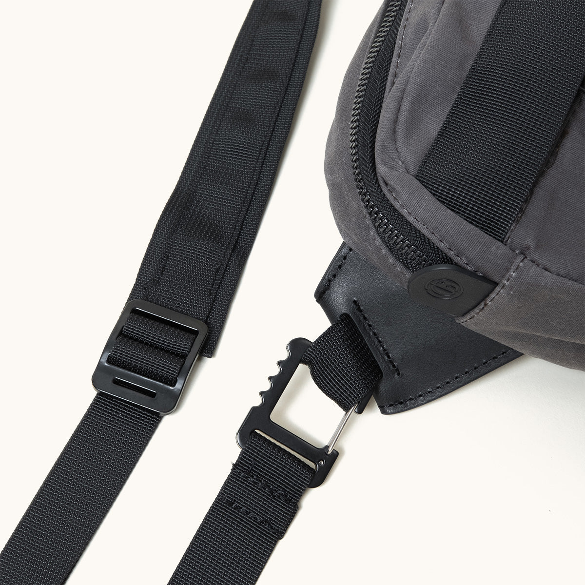 Slate grey waxed canvas crossbody pack with black nylon straps, black plastic hardware, and a debossed logo on the zipper.