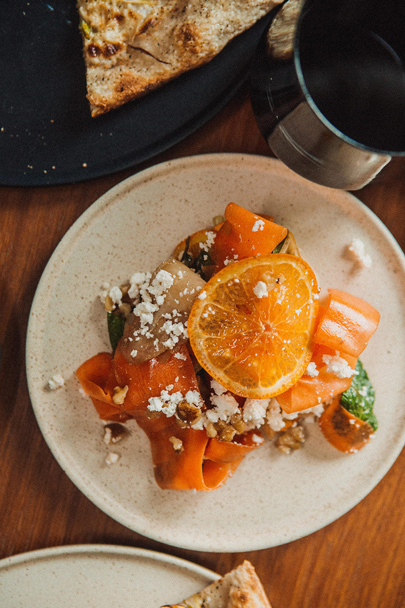 Speckled beige side plate topped with orange, cheese, and nuts, next to a pizza slice and black mug.