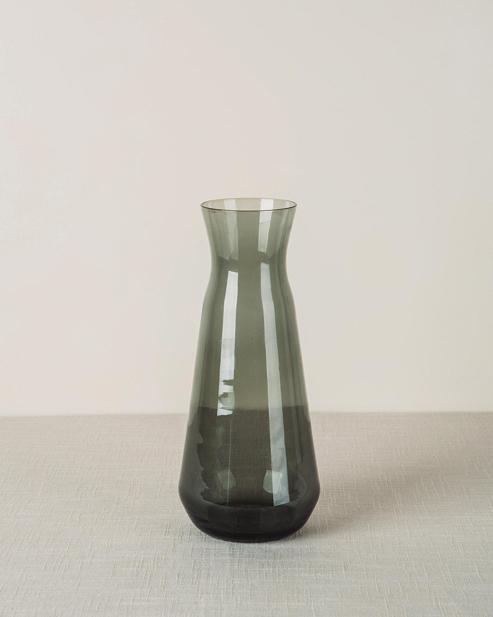 Dark grey glass carafe with a tapered shape on a light grey woven table surface.