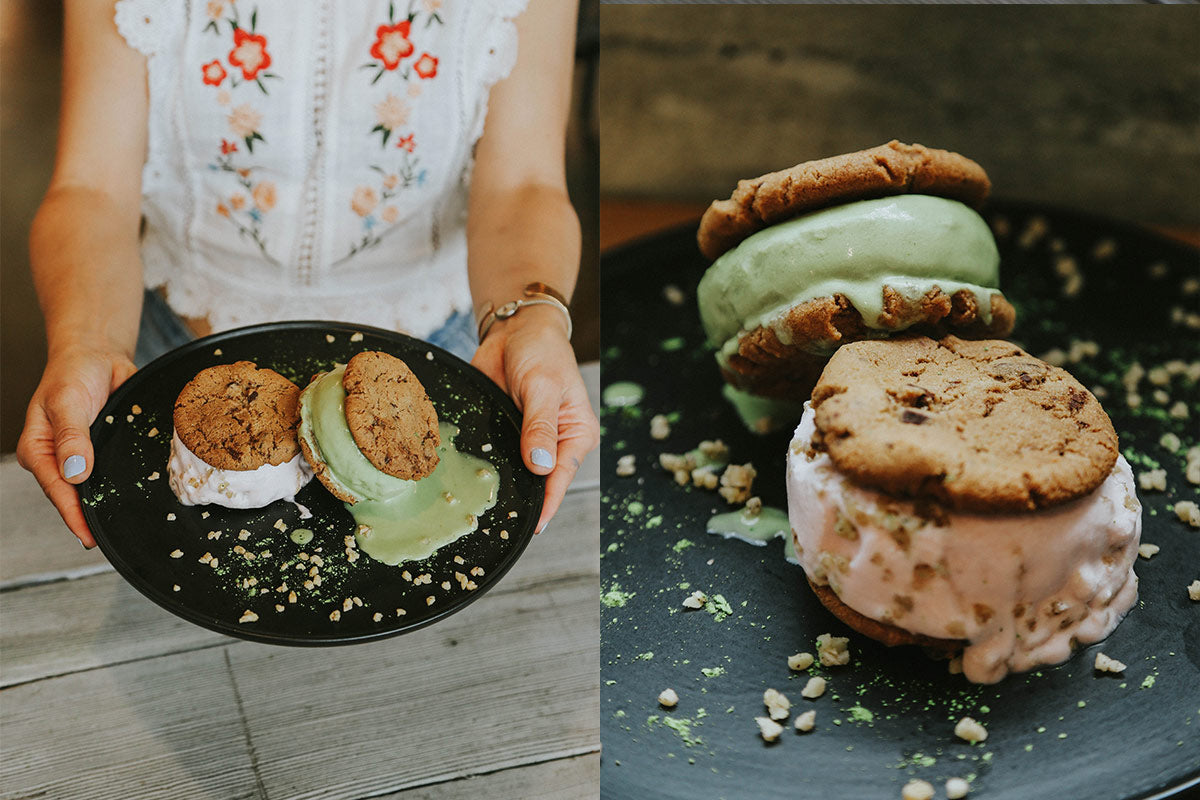 Black dinner plate held by hands, with two melting chocolate chip cookie ice cream sandwiches, green and pink, topped with nuts and green powder.