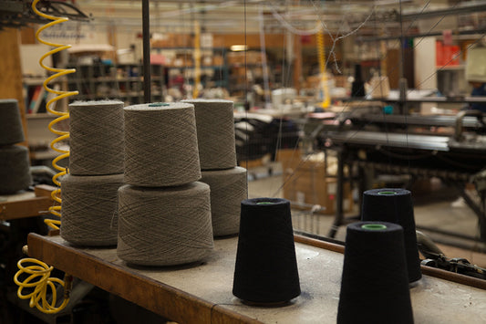 Stacks of gray and black yarn spools on a wooden table in a bustling knitting workshop, with machinery and yellow coiled hoses in the background.