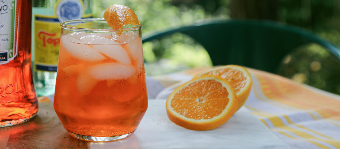 Vibrant orange spritz cocktail with ice and orange garnish, set on a sunny outdoor table with fresh orange slices and bottles nearby.