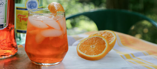 Vibrant orange spritz cocktail with ice and orange garnish, set on a sunny outdoor table with fresh orange slices and bottles nearby.