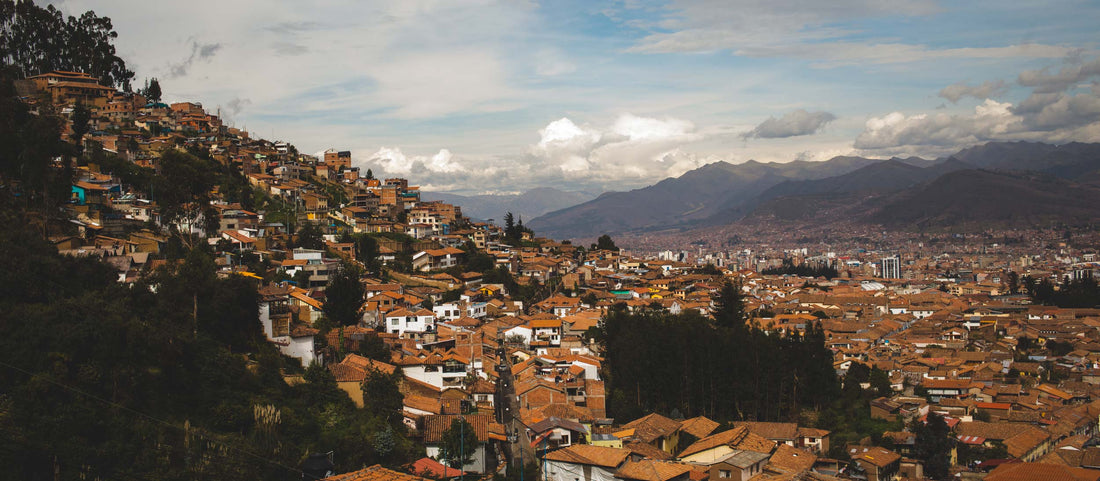Colorful terracotta rooftops cascade down hillsides in Cusco, Peru, with distant mountains under a cloudy sky.