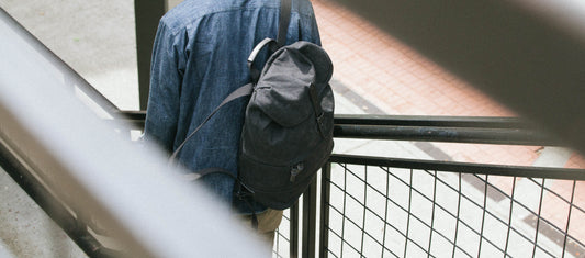 Person in denim shirt carries sleek black backpack on urban walkway, embodying minimalist travel style.