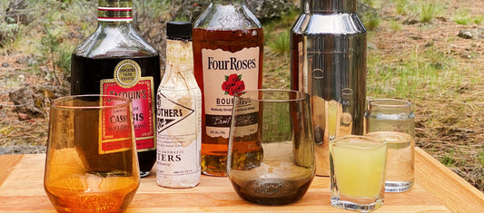 Wooden tray with bourbon, bitters, liqueur, shaker, and glasses outdoors—ready for a refreshing cocktail.