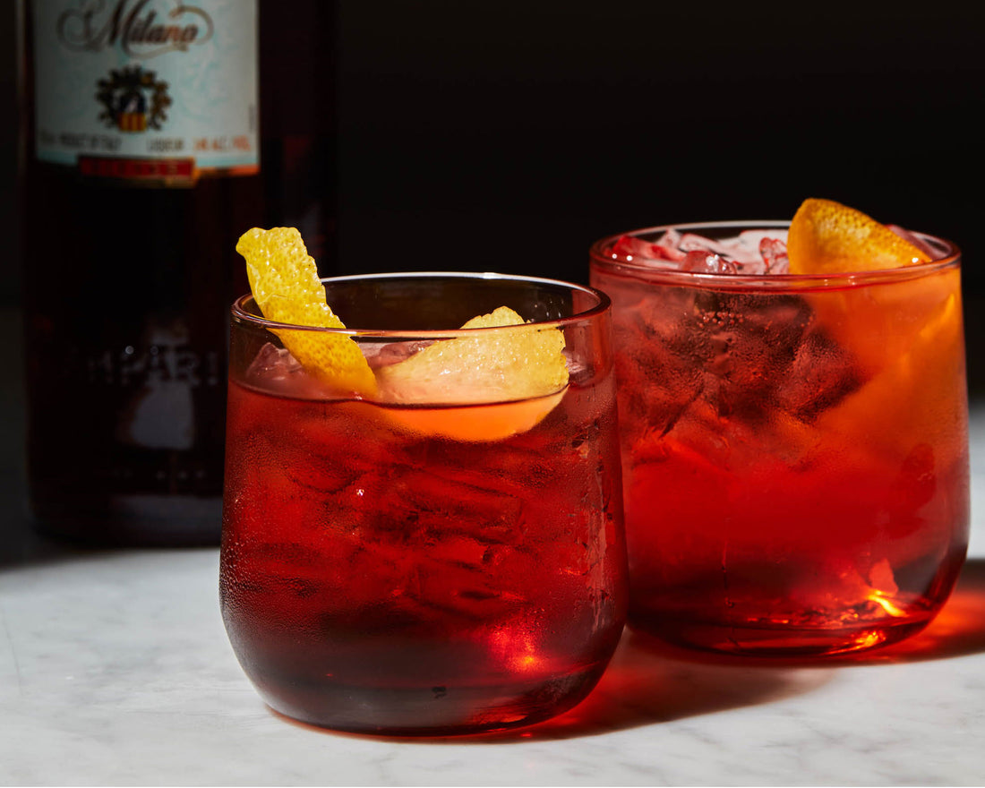 Two vibrant red Negronis with orange garnishes and ice, served in tumblers beside a bottle of Campari on a marble surface.