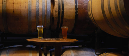 Two glasses of beer sit on a wooden table in a dimly lit basement lined with large wooden barrels.