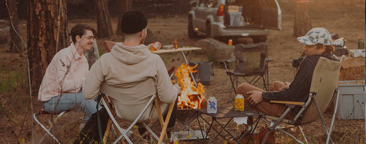 Three friends relax around a campfire in a forest, laughing and sharing drinks, embodying simple, meaningful adventure.