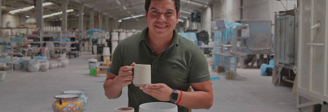 Smiling craftsman holds handmade ceramic mug in bustling Mexican pottery workshop, showcasing artisan pride.