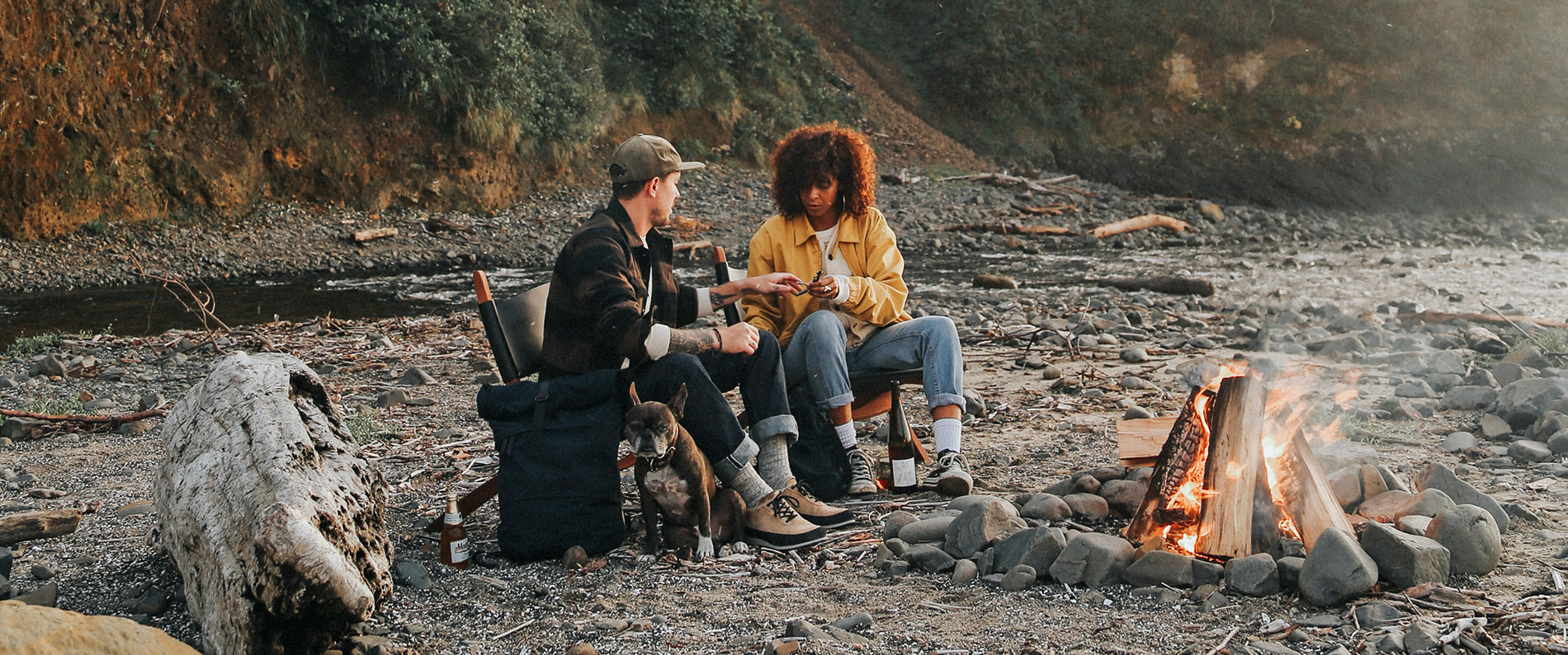 Couple in rugged outdoor wear relax by campfire with dog, enjoying nature in earthy tones and cozy vibes.