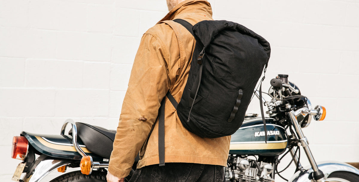 Man in tan jacket with black backpack stands beside vintage Kawasaki motorcycle, urban adventure style.