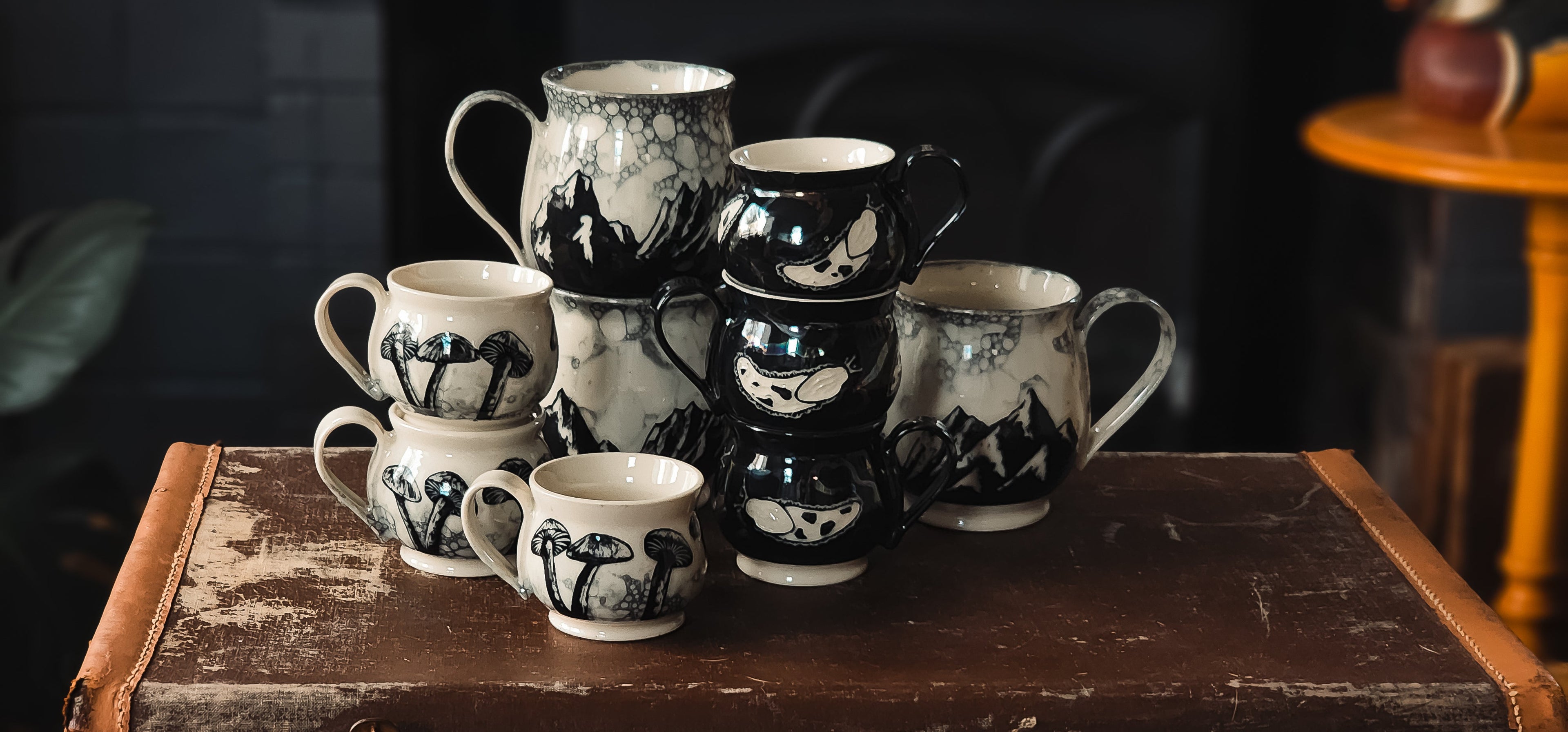 Hand-painted ceramic mugs and pitcher with black-and-white mountain, mushroom, and abstract patterns, styled on vintage trunk.