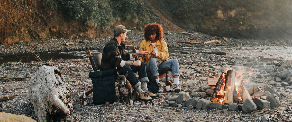 Couple in rugged outdoor wear relax by campfire with dog, enjoying nature in earthy tones and cozy vibes.