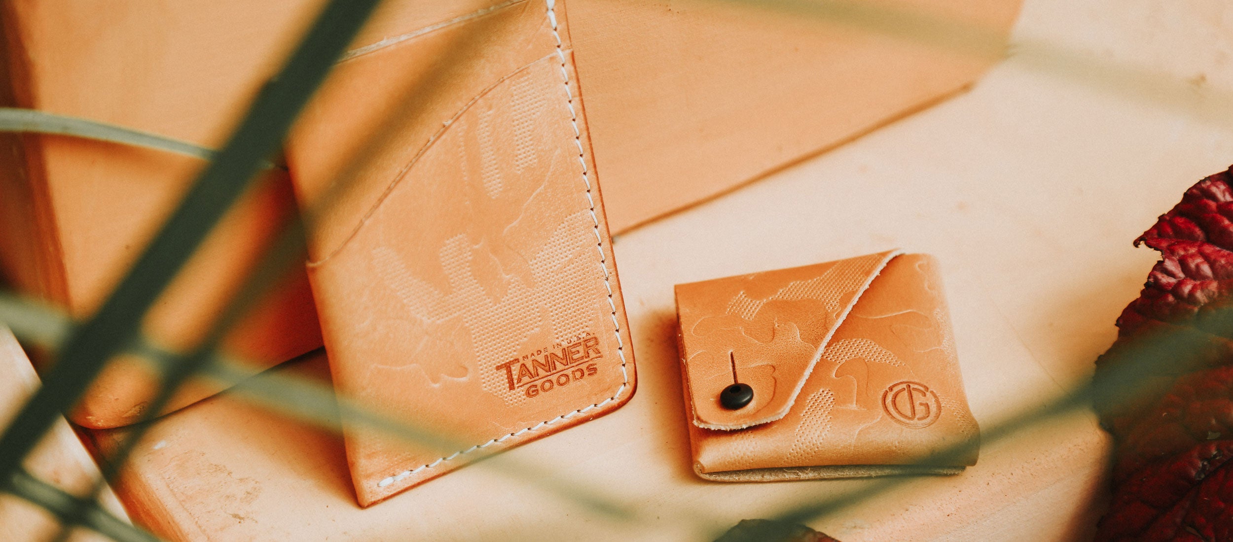 Tan leather wallets with embossed camouflage pattern, white stitching, and Tanner Goods branding, styled with natural light and foliage.