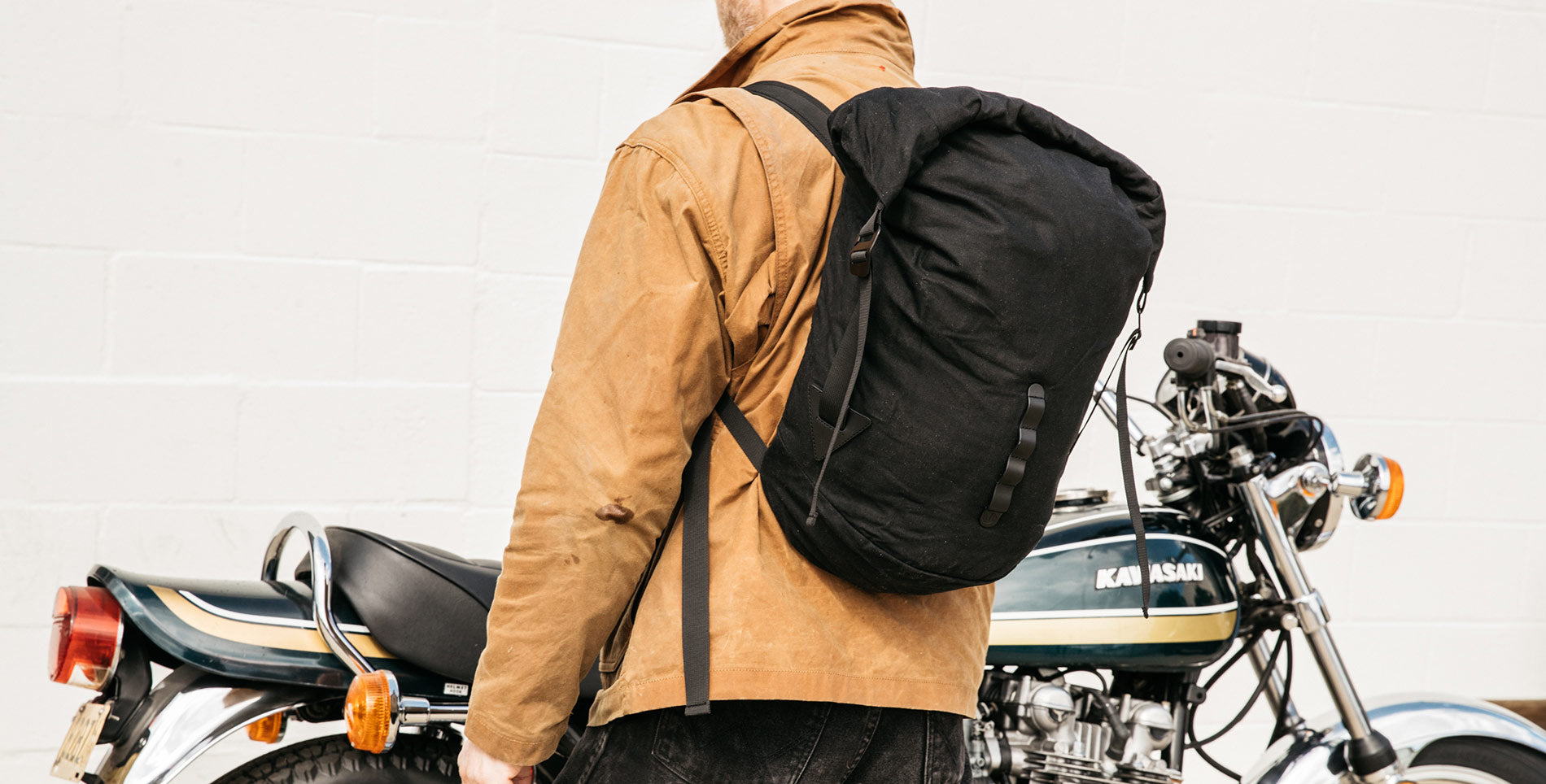 Man in tan jacket with black backpack stands beside vintage Kawasaki motorcycle, urban adventure style.