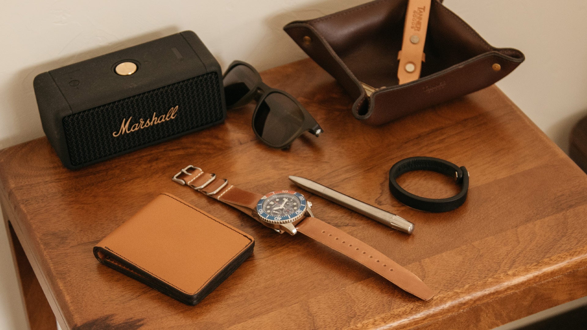 Rich wood table displays premium leather wallet, watch, pen, speaker, and sunglasses—classic, functional, stylish essentials for the modern student.