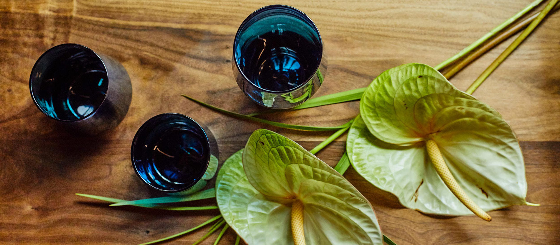 Deep blue hand-blown glass tumblers on warm wood with lush green anthuriums, evoking natural elegance and artisan craftsmanship.