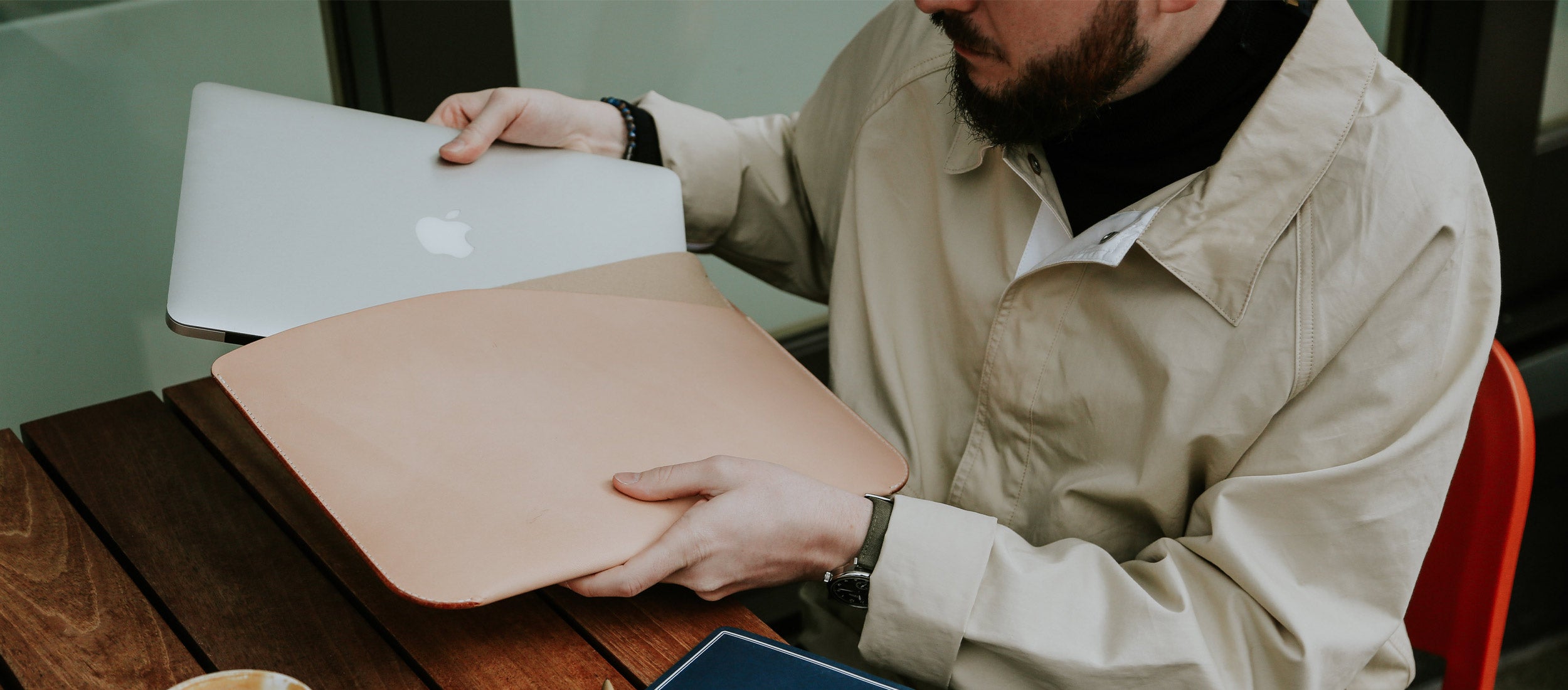Man placing MacBook into tan leather sleeve at wooden desk, minimalist office essentials in neutral tones.