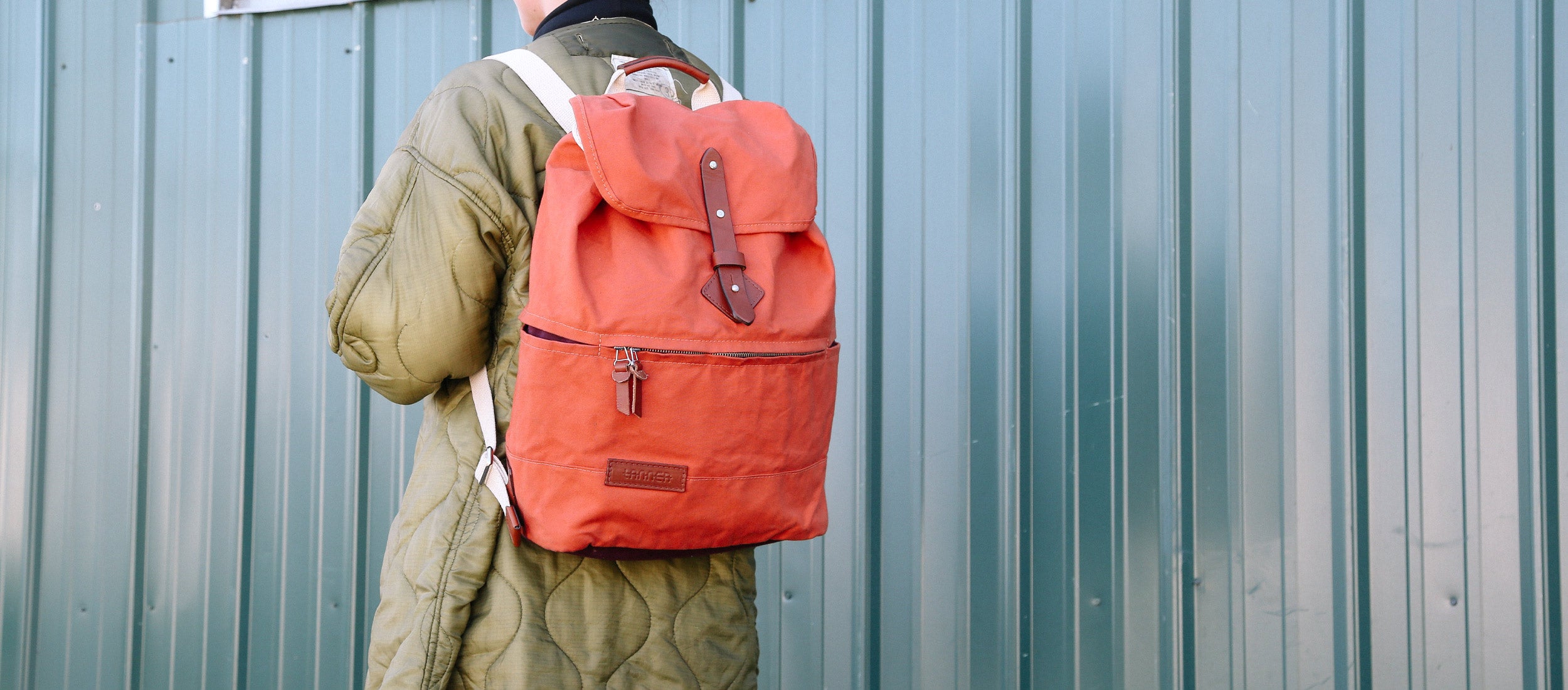 Vibrant coral backpack with leather accents worn by person in olive quilted jacket against teal corrugated wall, urban adventure style.