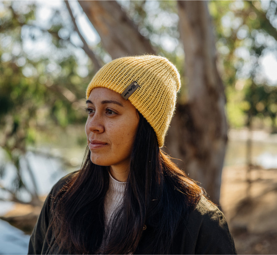Golden knit beanie with subtle logo, worn by woman in natural light, earthy tones, outdoor lifestyle.