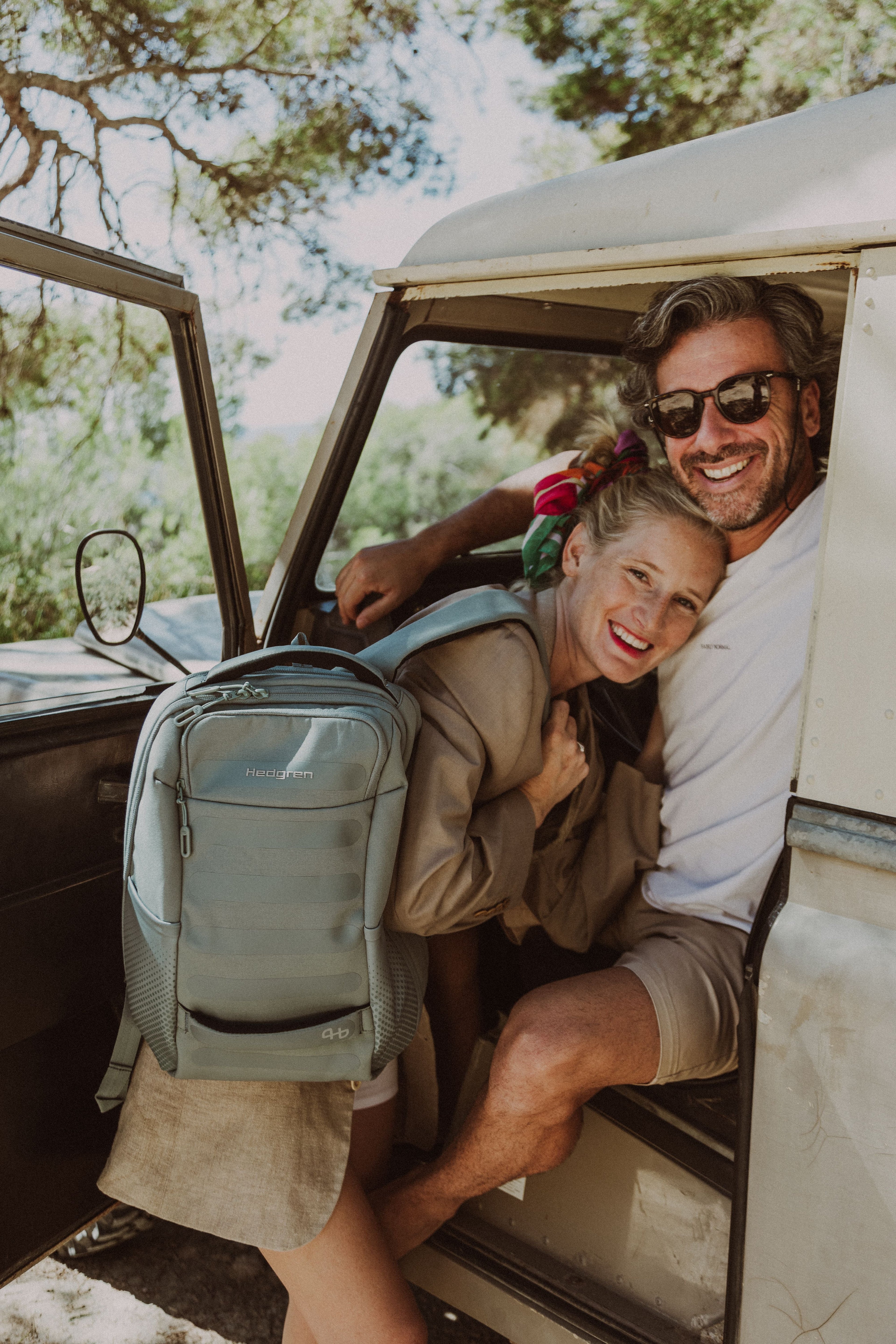 Hedgren backpack in sage green, worn by smiling couple in vintage car, embodying adventure and modern travel style.
