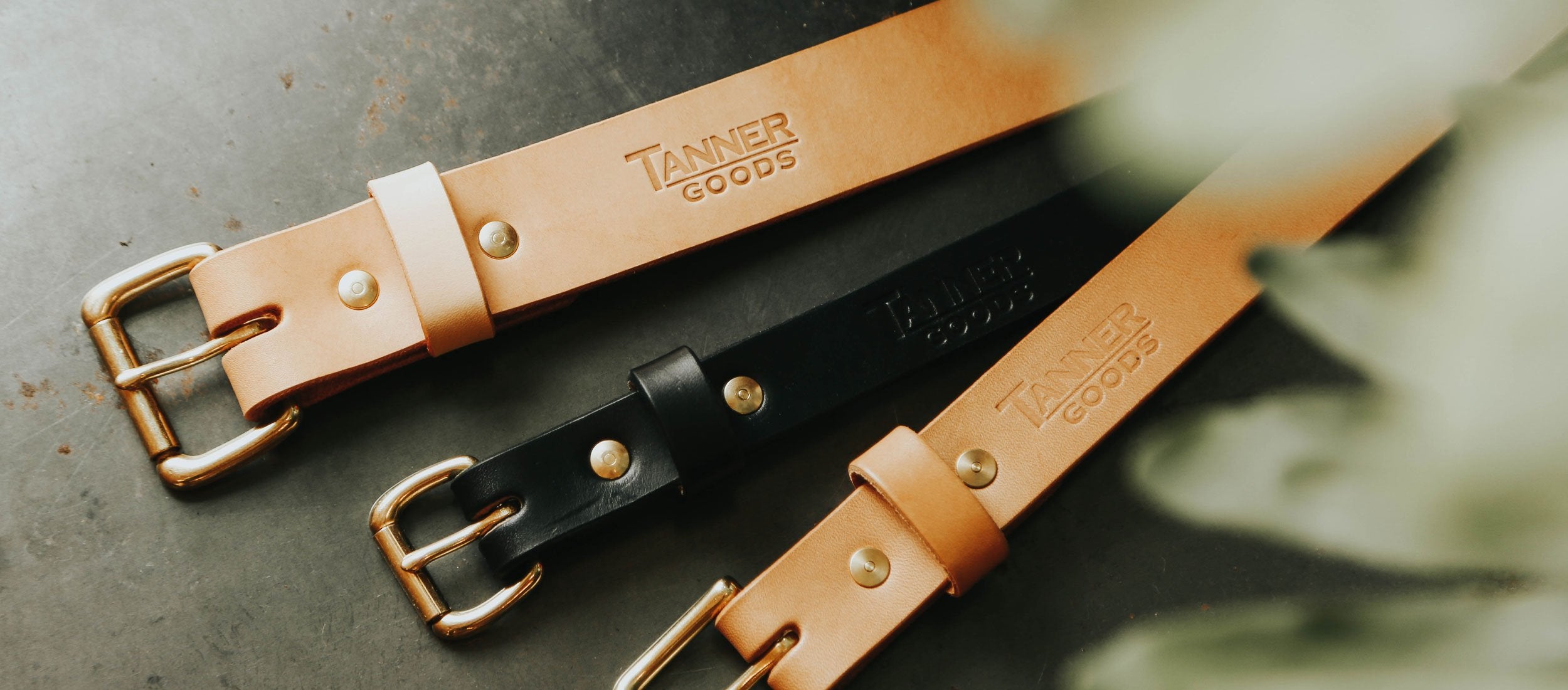 Rich tan and black leather belts with brass buckles, embossed "Tanner Goods" — timeless, durable gifts for the modern gentleman.