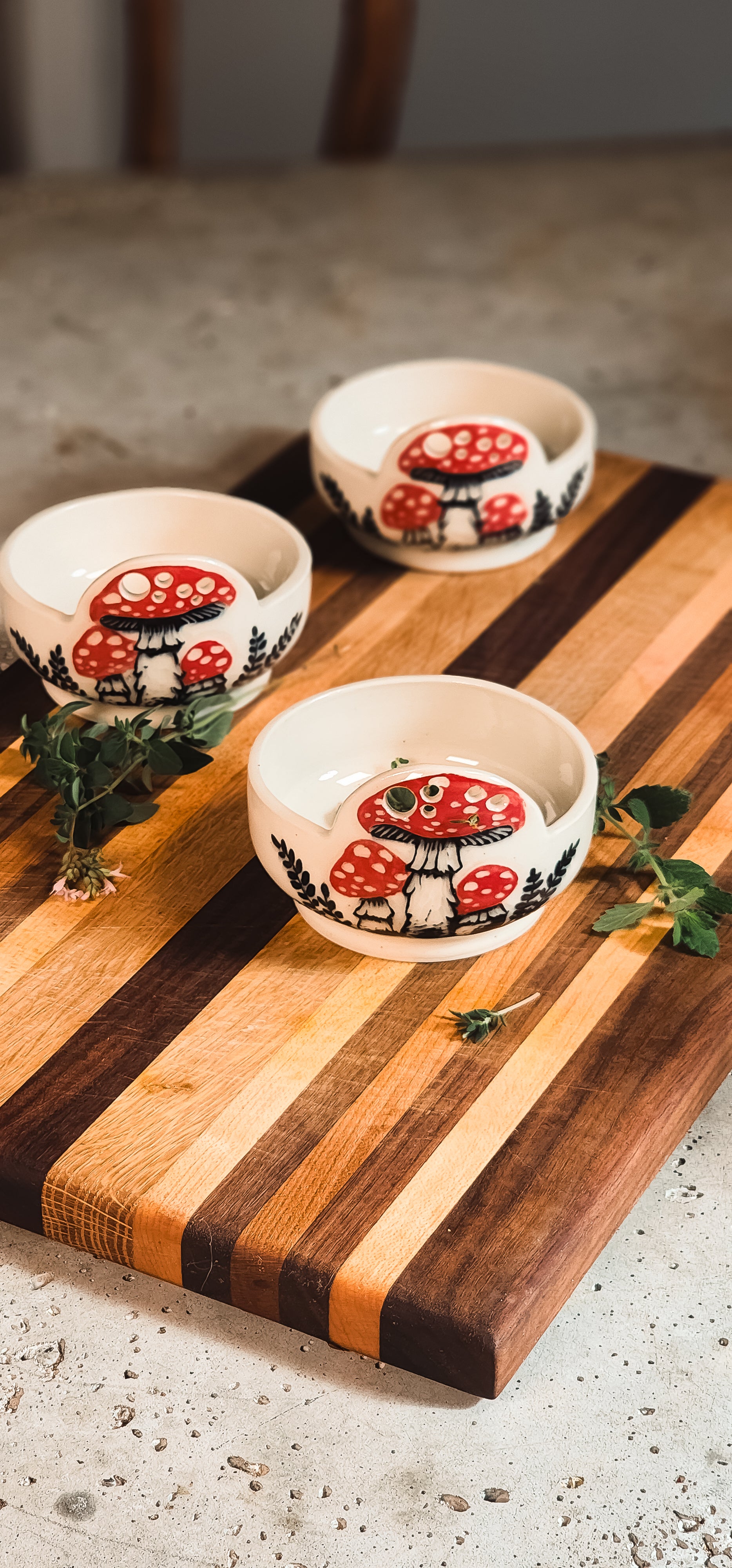 White ceramic herb puller with red and white mushroom design, displayed on a striped wooden cutting board with fresh green herbs.