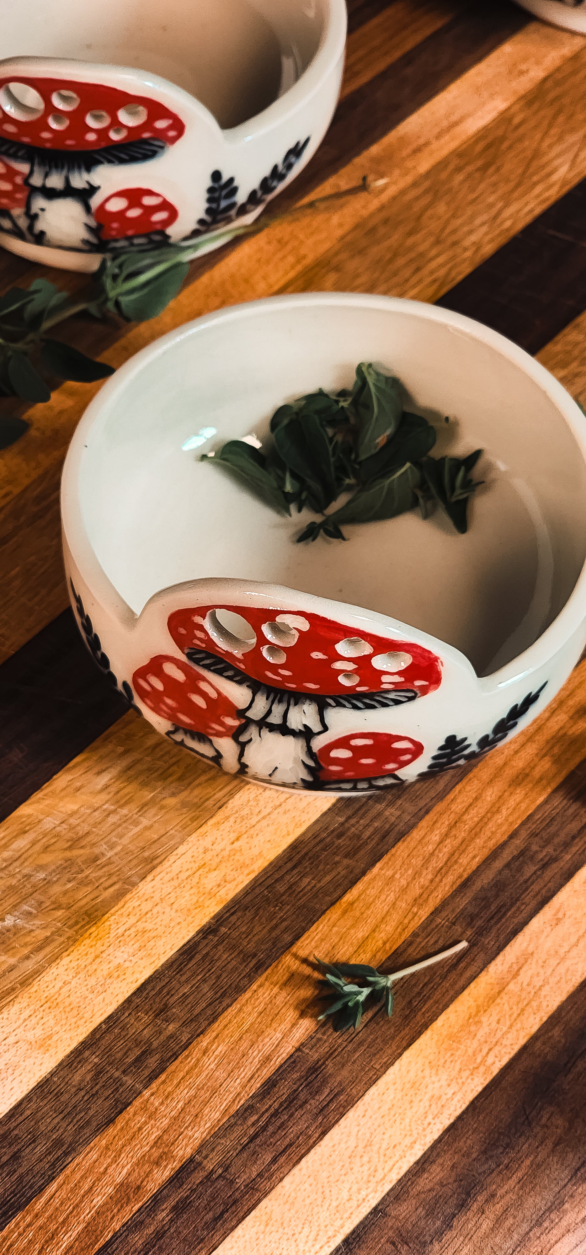 Two white ceramic herb pullers with red mushroom designs, one filled with fresh herbs, on a striped wooden board.
