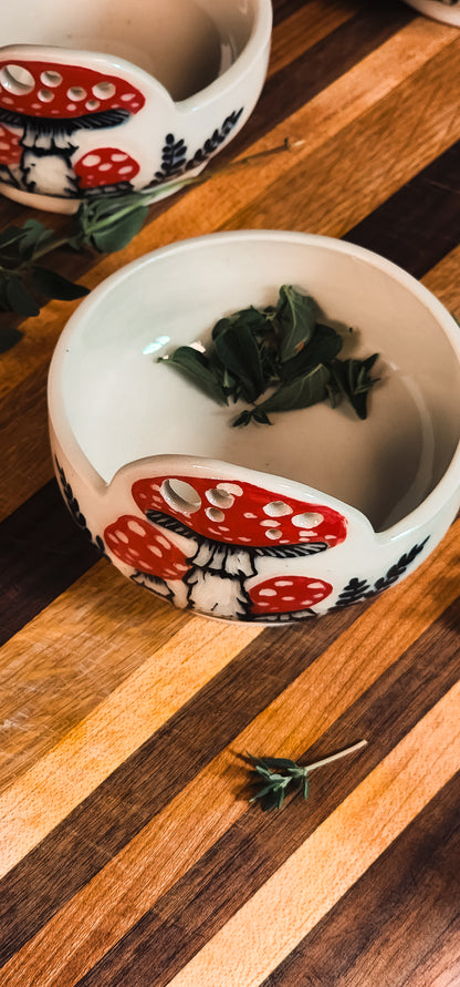Two white ceramic herb pullers with red mushroom designs, one filled with fresh herbs, on a striped wooden board.