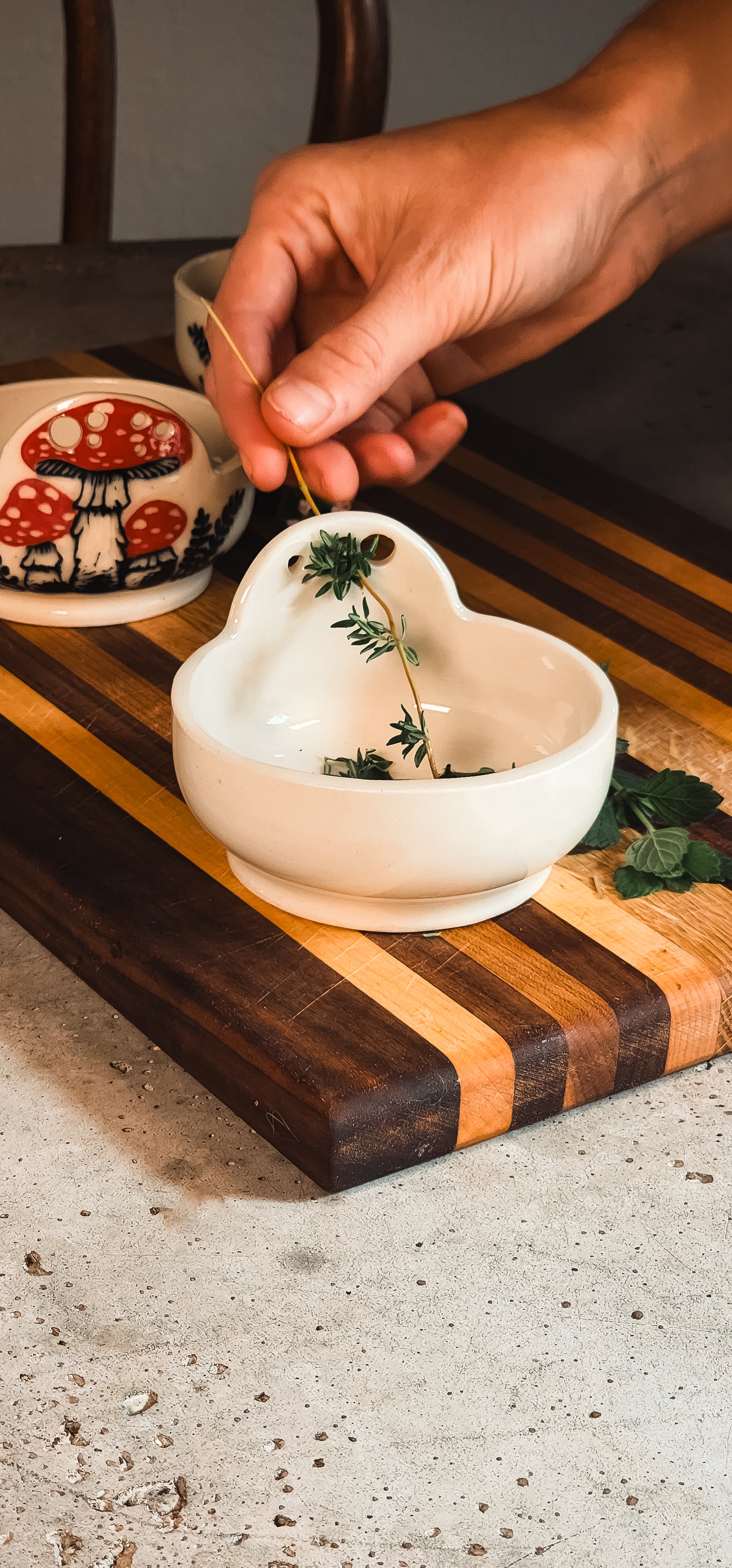 White ceramic heart-shaped herb puller holding thyme sprigs, with a hand pulling a stem through a hole on a striped cutting board.