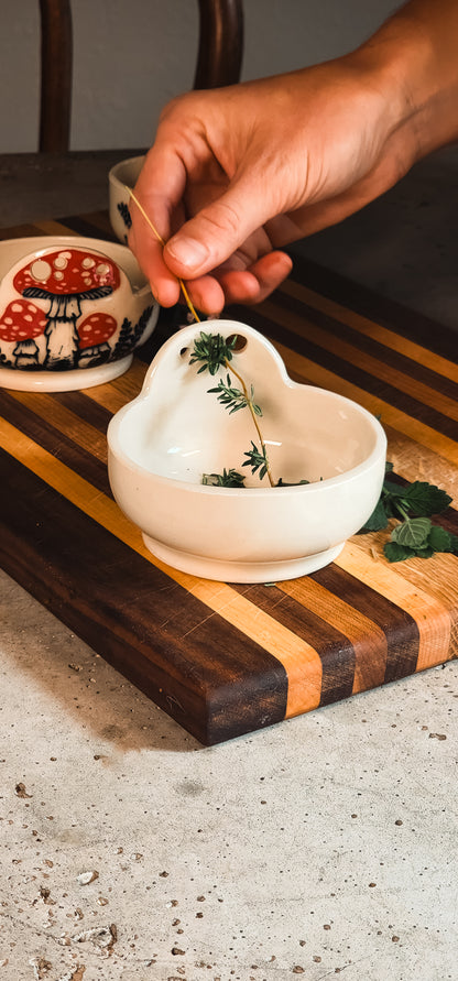 White ceramic heart-shaped herb puller holding thyme sprigs, with a hand pulling a stem through a hole on a striped cutting board.