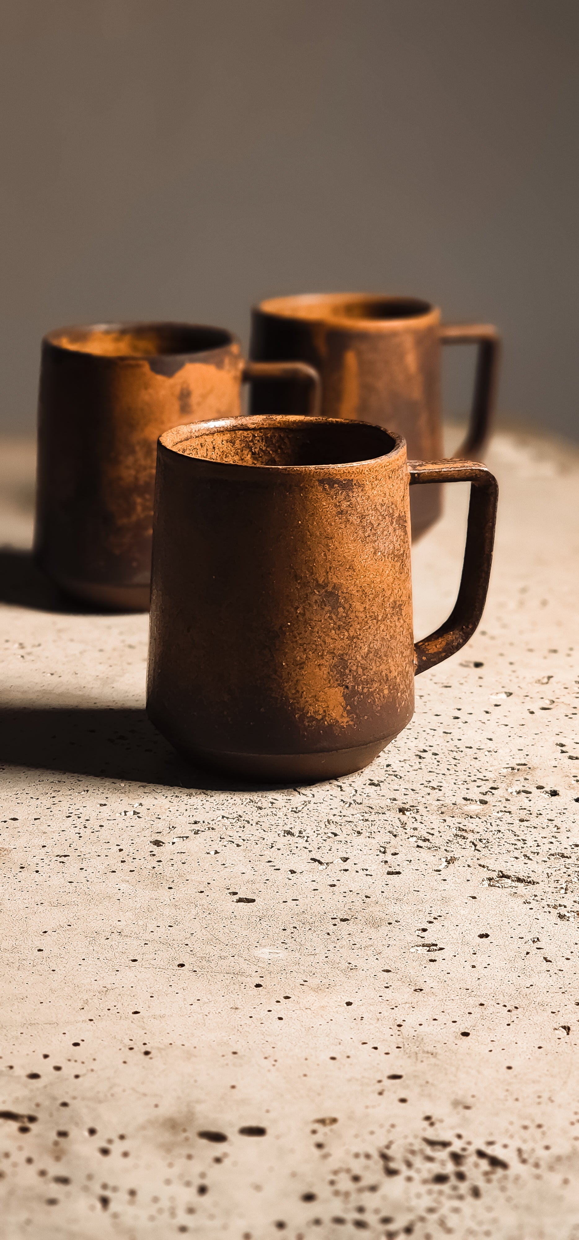 Rustic distressed brown standard mug with a handle, sitting on a speckled concrete surface with two blurred mugs behind.