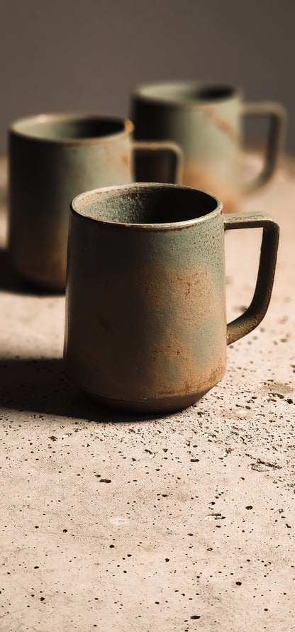 Earthy green and brown ceramic standard mug with a rustic, textured finish on a speckled surface.