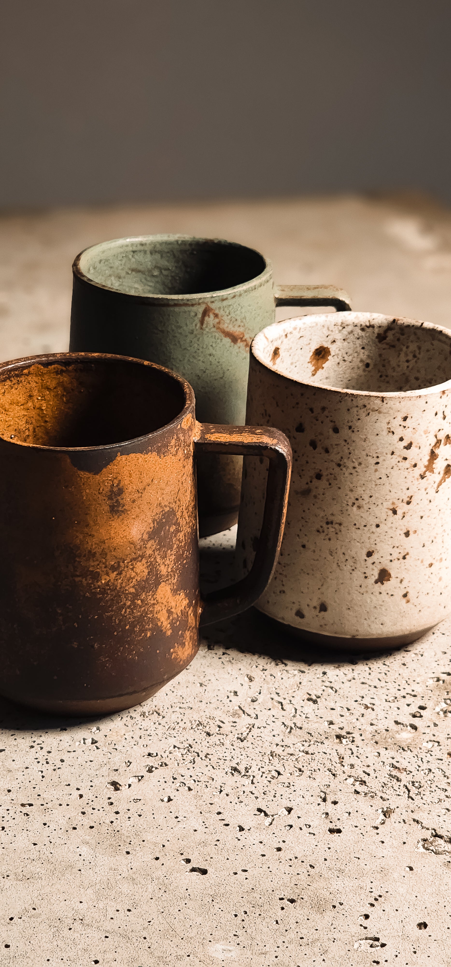 Three standard mugs of rustic ceramic, in brown, green, and speckled beige with textured glazes on a concrete surface.