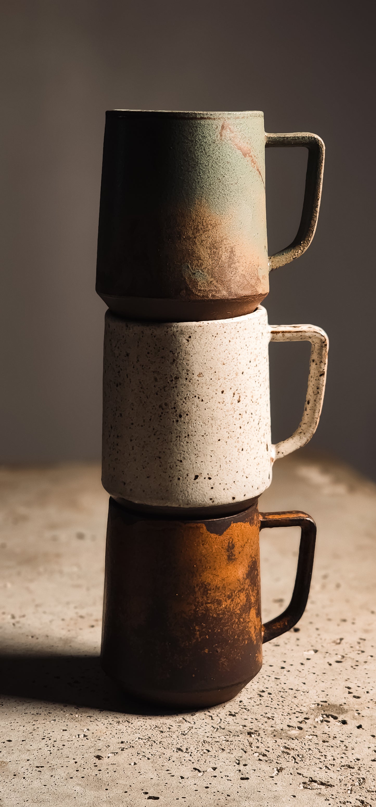Three stacked standard mugs with textured surfaces: green, white speckled, and rusty brown.