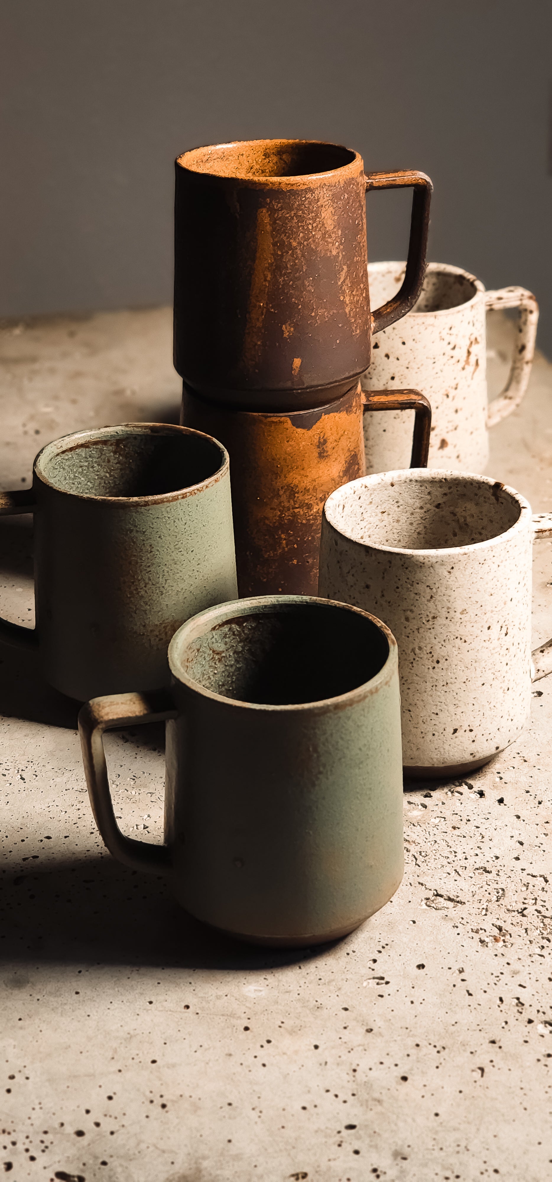 Rustic standard mugs in speckled brown, beige, and green ceramic, some stacked, displayed on a textured surface.