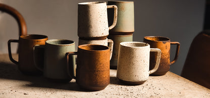 Assorted ceramic standard mugs in speckled cream, light green, and rustic brown, some stacked, on a textured surface.