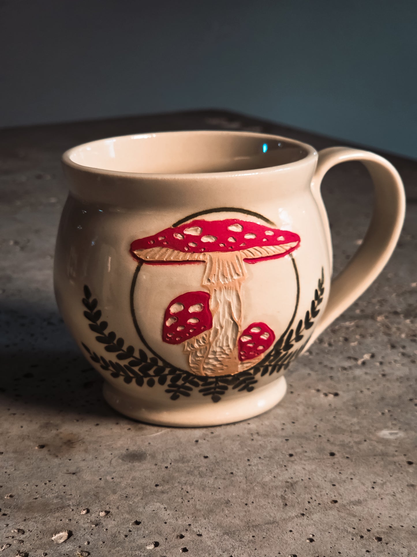 Amanita mushroom mug with raised red and white mushroom design and dark leaf patterns on a cream background.