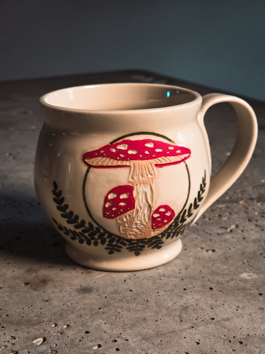 Amanita mushroom mug with raised red and white mushroom design and dark leaf patterns on a cream background.