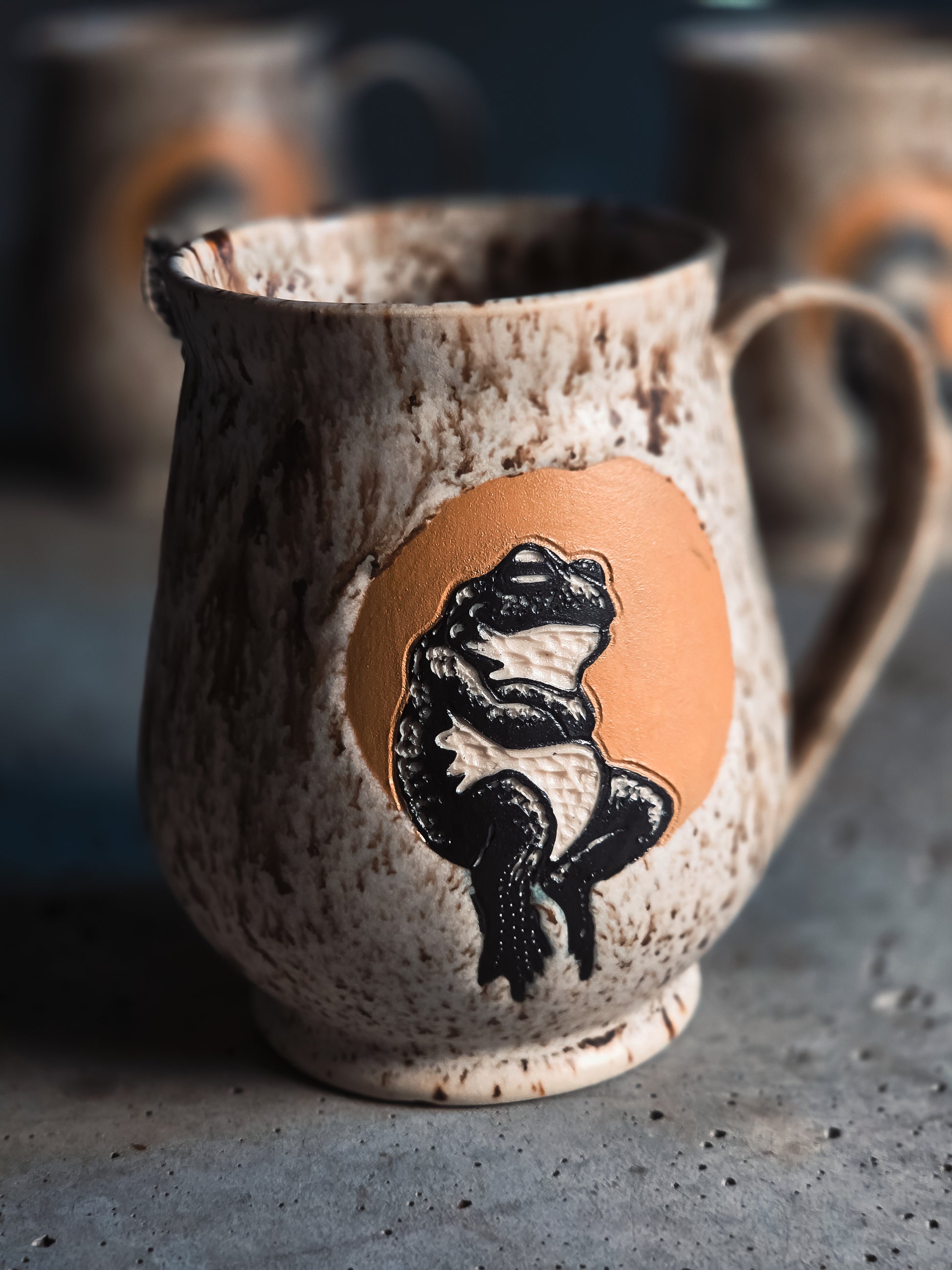 Speckled ceramic pitcher featuring a black frog design on an orange circle, on a textured grey surface.