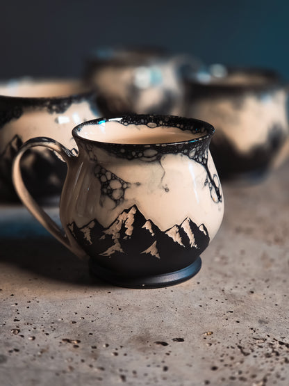 Cream and black ceramic mug with a black bubble patterned rim and mountain silhouettes, on a concrete surface.