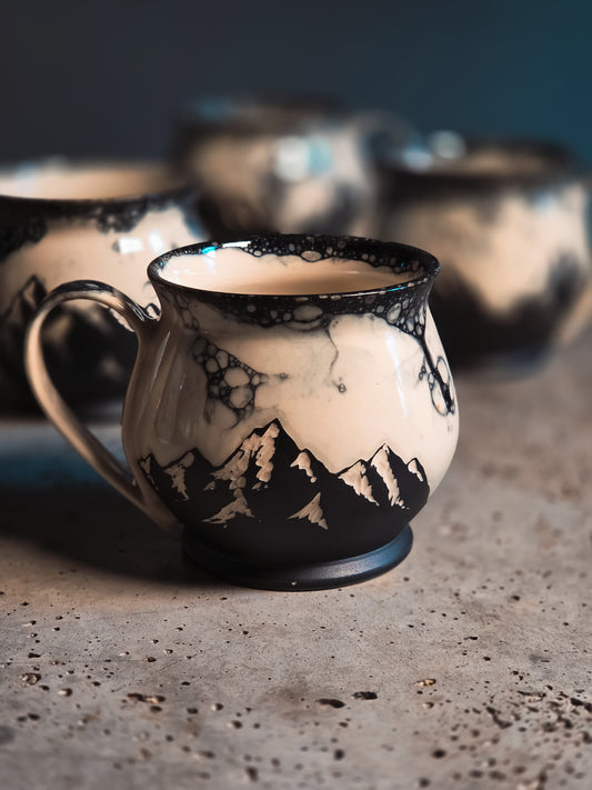 Cream and black ceramic mug with a black bubble patterned rim and mountain silhouettes, on a concrete surface.
