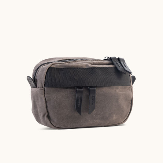 Slate grey waxed canvas crossbody mini bag with a top zipper, two black pull tabs, and a black front strap, angled.