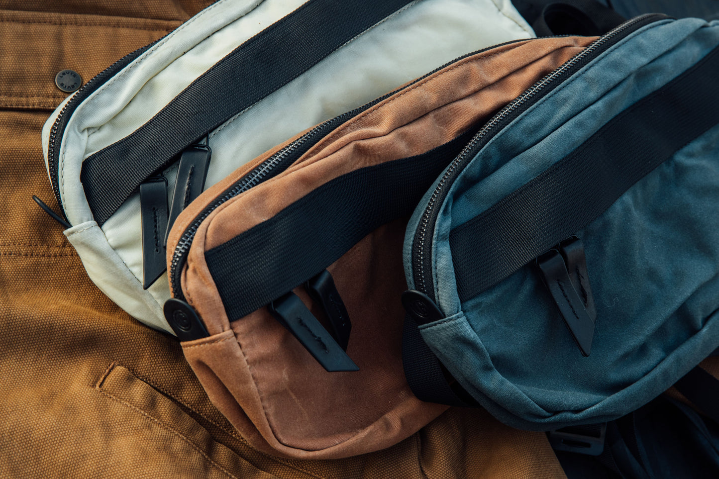 Frost waxed canvas Canyon Crossbody Pack, prominently stacked with burnt orange and dark teal packs, featuring black zippers and straps.