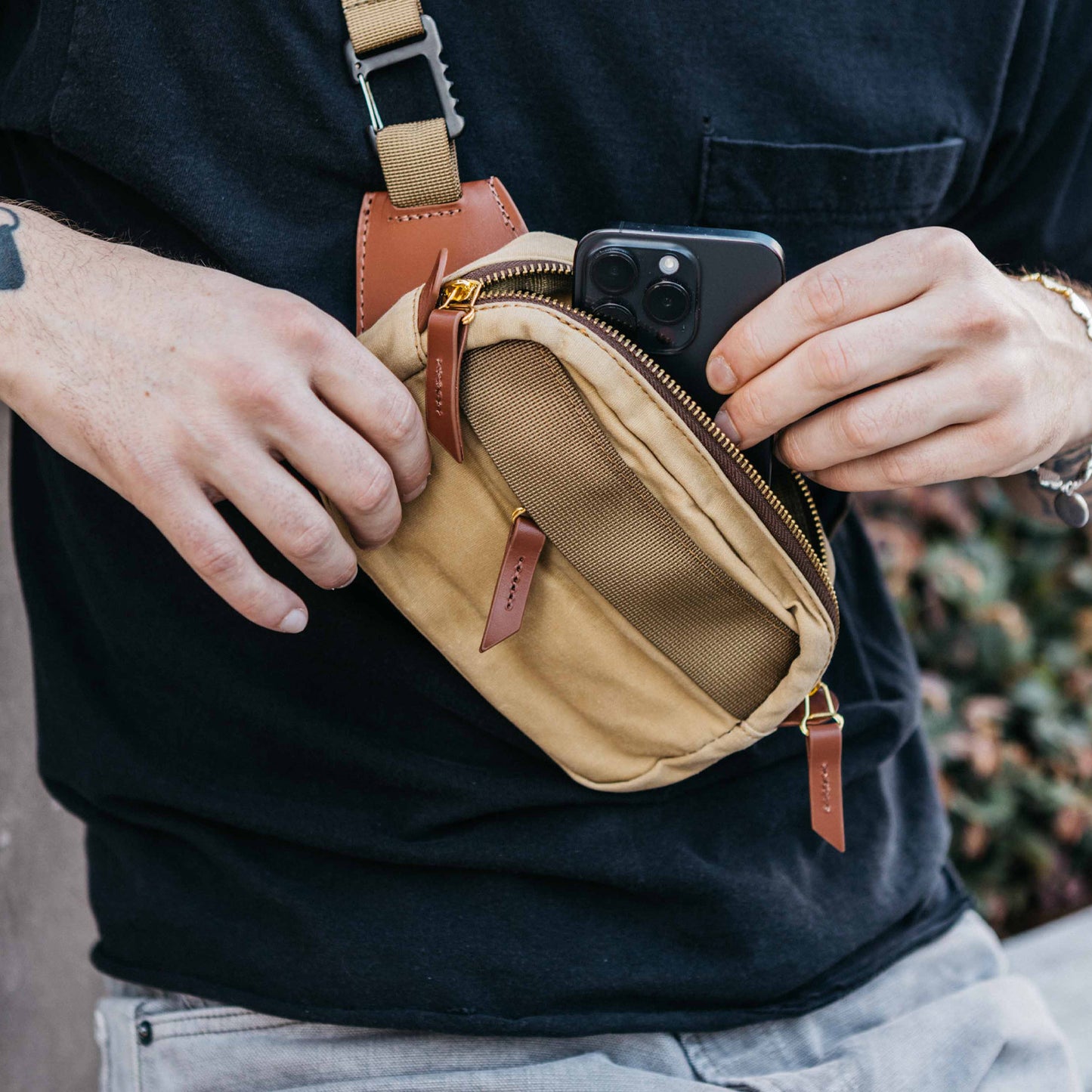 Tan crossbody mini bag with a visible zipper, brown leather pull tabs, and mesh front pocket, held by a person inserting a phone.