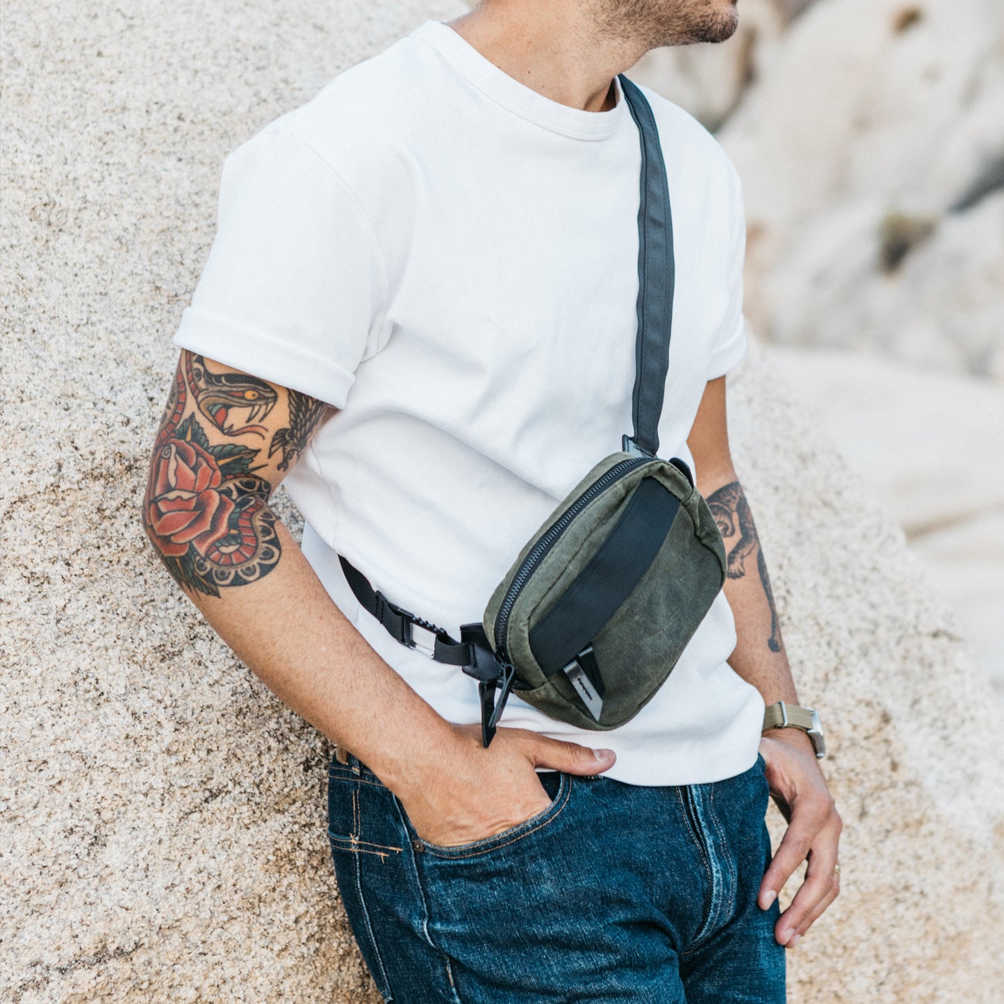 Olive green waxed canvas crossbody mini bag with black strap details, worn by a man in a white t-shirt and jeans, leaning on a rock.
