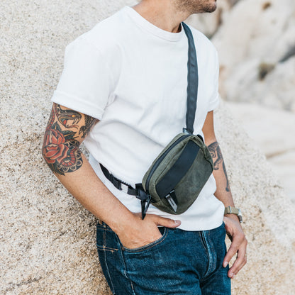 Olive green waxed canvas crossbody mini bag with black strap details, worn by a man in a white t-shirt and jeans, leaning on a rock.