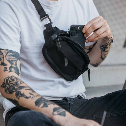 Slate Grey Waxed Canvas Canyon Crossbody Mini, worn across the chest by a person putting a black smartphone into its zippered compartment.